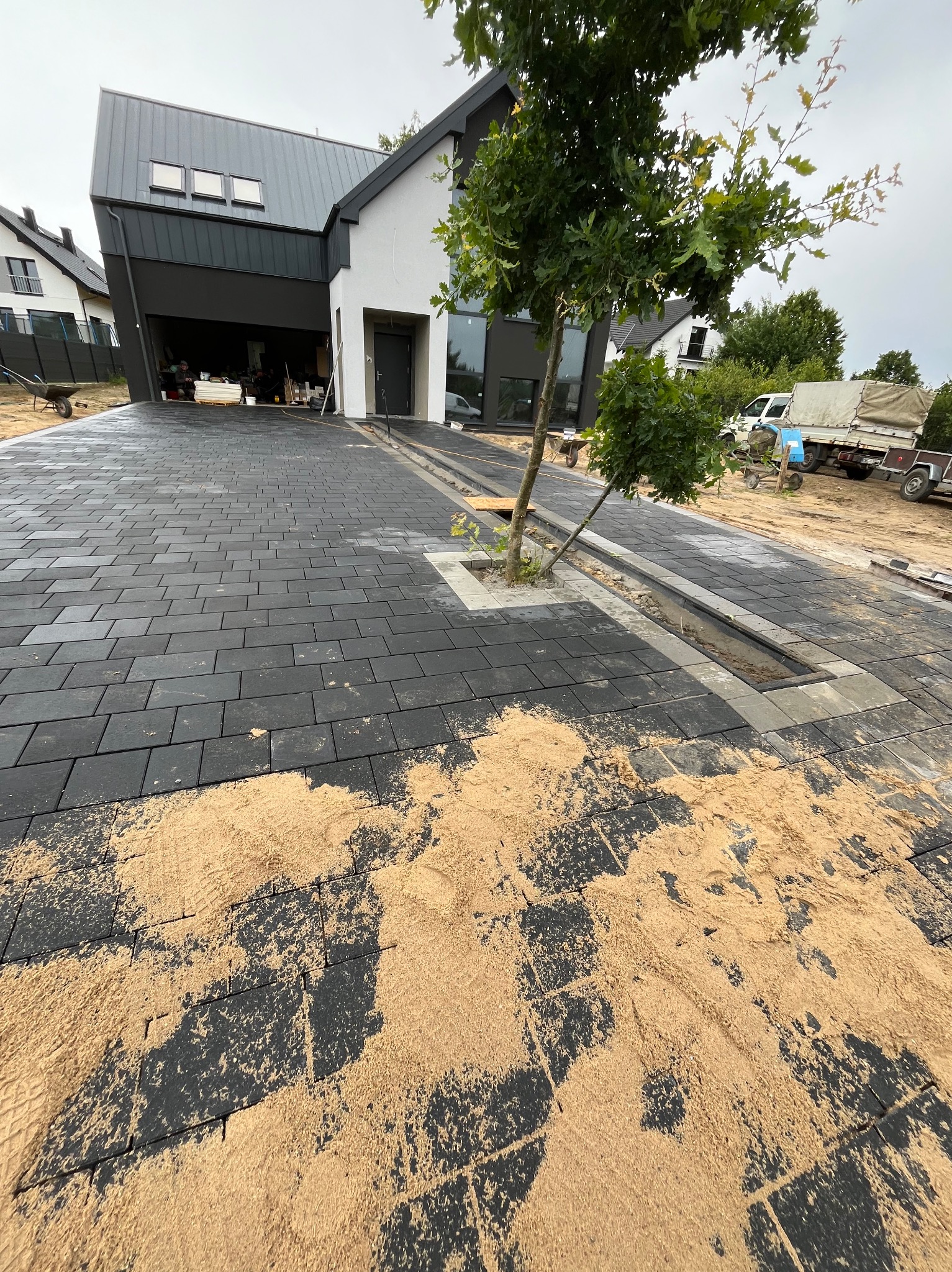 Nowoczesny podjazd z ciemnej kostki brukowej w trakcie układania, z widocznym piaskiem do wypełniania szczelin, z wbudowanym odpływem liniowym i miejscem na drzewa, na tle nowoczesnego domu z garażem.