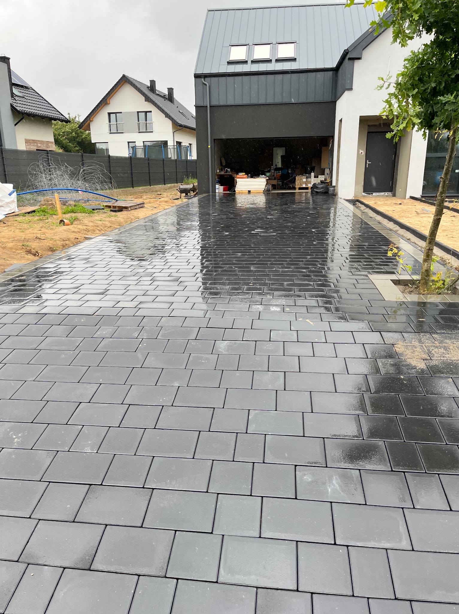 Świeżo ułożona, mokra kostka brukowa w kolorze grafitowym prowadząca do nowoczesnego domu z otwartym garażem; widoczne przygotowane podłoże po bokach i drzewko z obrzeżem.