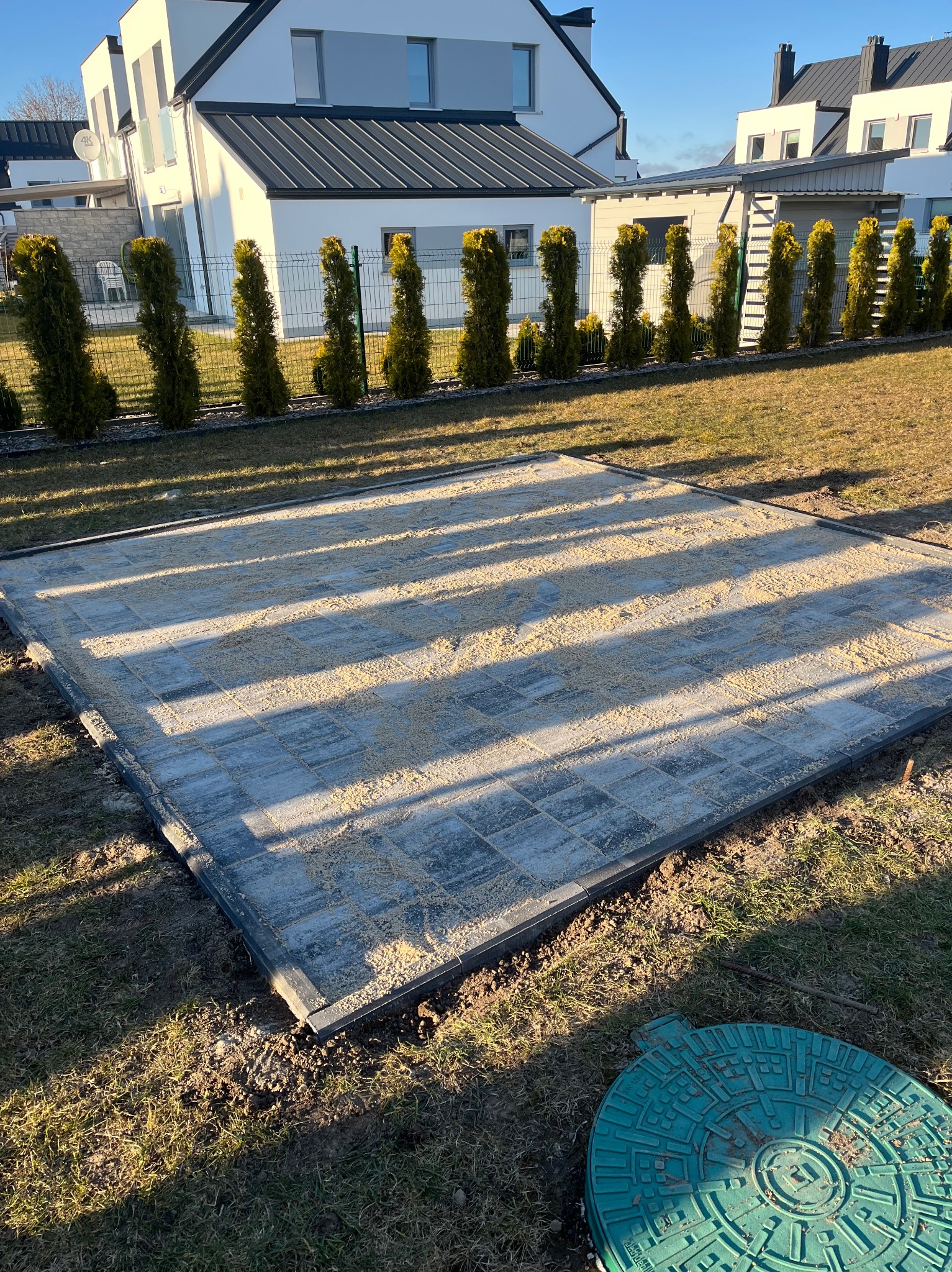 Przygotowany teren pod kostkę brukową z podsypką piaskową, widoczny fragment trawnika i studzienka kanalizacyjna, w tle rząd tuj i dom jednorodzinny.