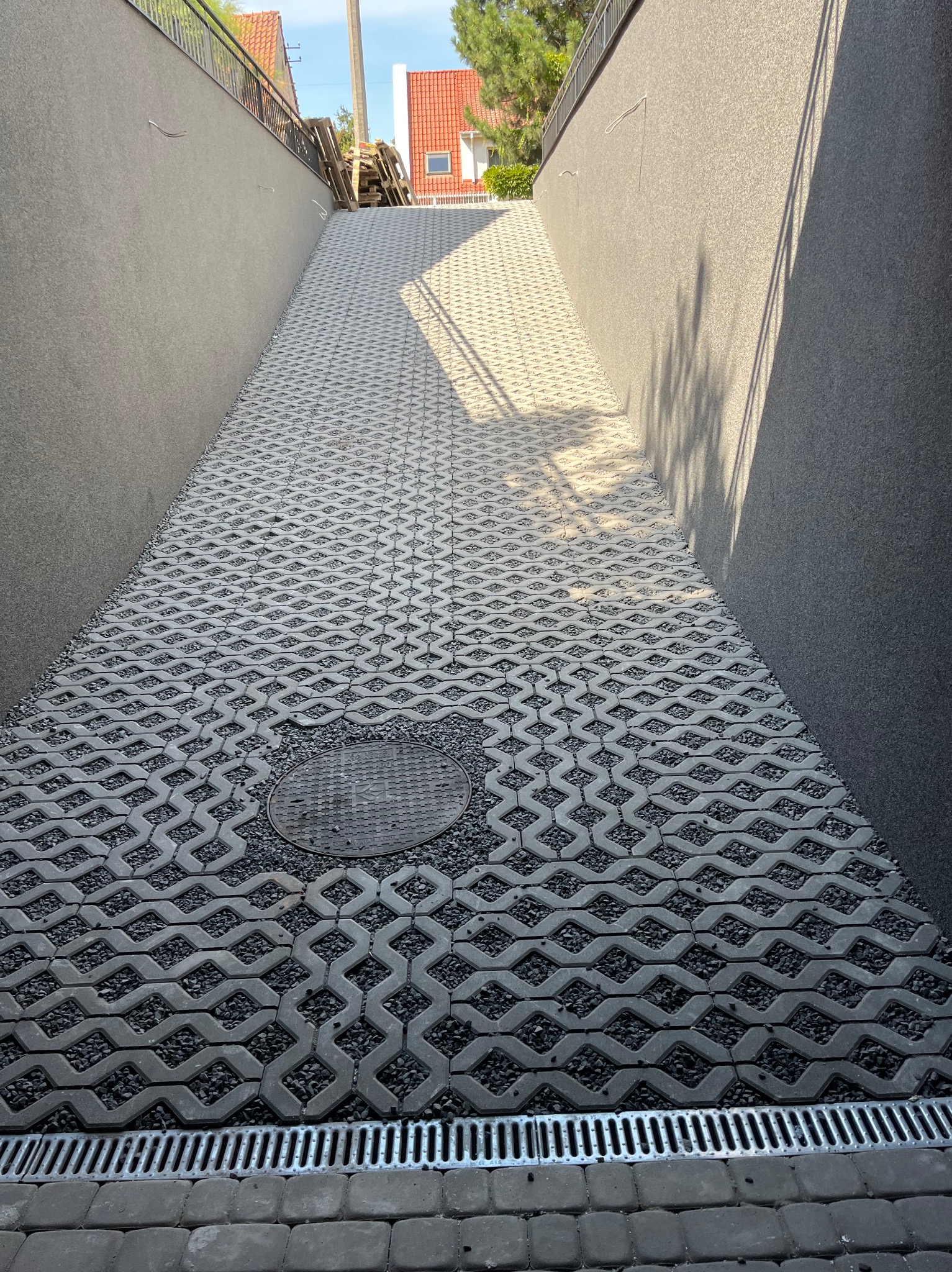 Podjazd wyłożony ażurową kostką betonową z wypełnieniem z drobnego kamienia, widoczna studzienka kanalizacyjna i metalowy odpływ liniowy, wysokie ściany po bokach podjazdu.