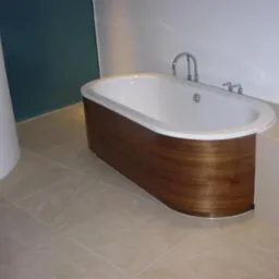 Nowoczesna wanna owalna z obudową z drewna orzechowego, chromowana bateria, beżowe płytki podłogowe i fragment ściany w kolorze petrol w tle.