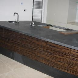 MODEN s.c. - Nowoczesna wyspa kuchenna z ciemnym blatem i frontami z egzotycznego drewna. Umywalka wpuszczona w blat, widoczne elementy instalacji. Minimalistyczny design.