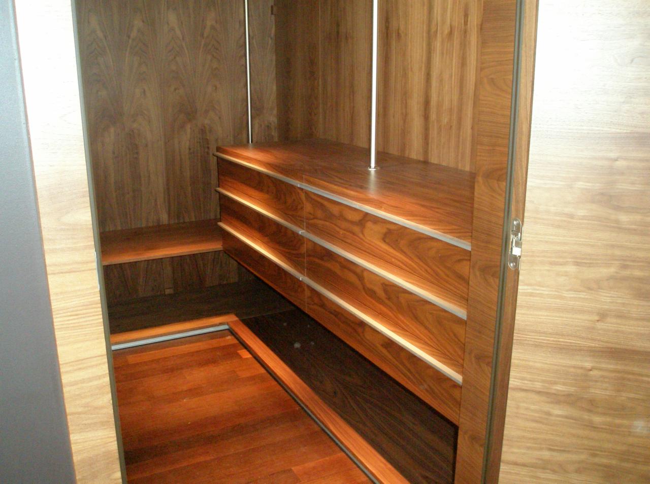 Wnętrze garderoby z drewnianymi elementami: szuflady z metalowymi akcentami, półki i podłoga z drewna o ciepłym odcieniu, widoczne elementy oświetlenia.