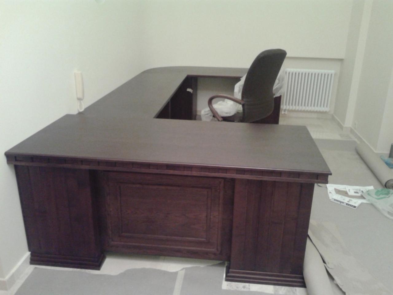 Ciemnobrązowe, drewniane biurko narożne z krzesłem biurowym w tle, na podłodze zabezpieczenia. Widoczny fragment ściany z telefonem.