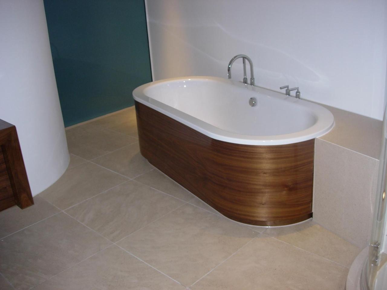 Nowoczesna wanna owalna z obudową z drewna orzechowego, chromowana bateria, beżowe płytki podłogowe i fragment ściany w kolorze petrol w tle.