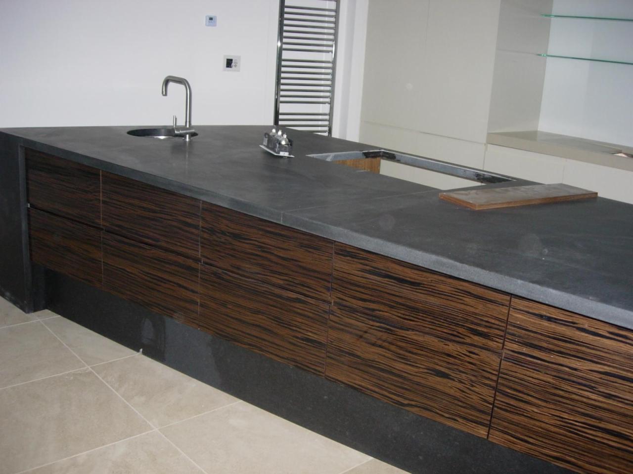 Nowoczesna wyspa kuchenna z ciemnym blatem i frontami z egzotycznego drewna. Umywalka wpuszczona w blat, widoczne elementy instalacji. Minimalistyczny design.