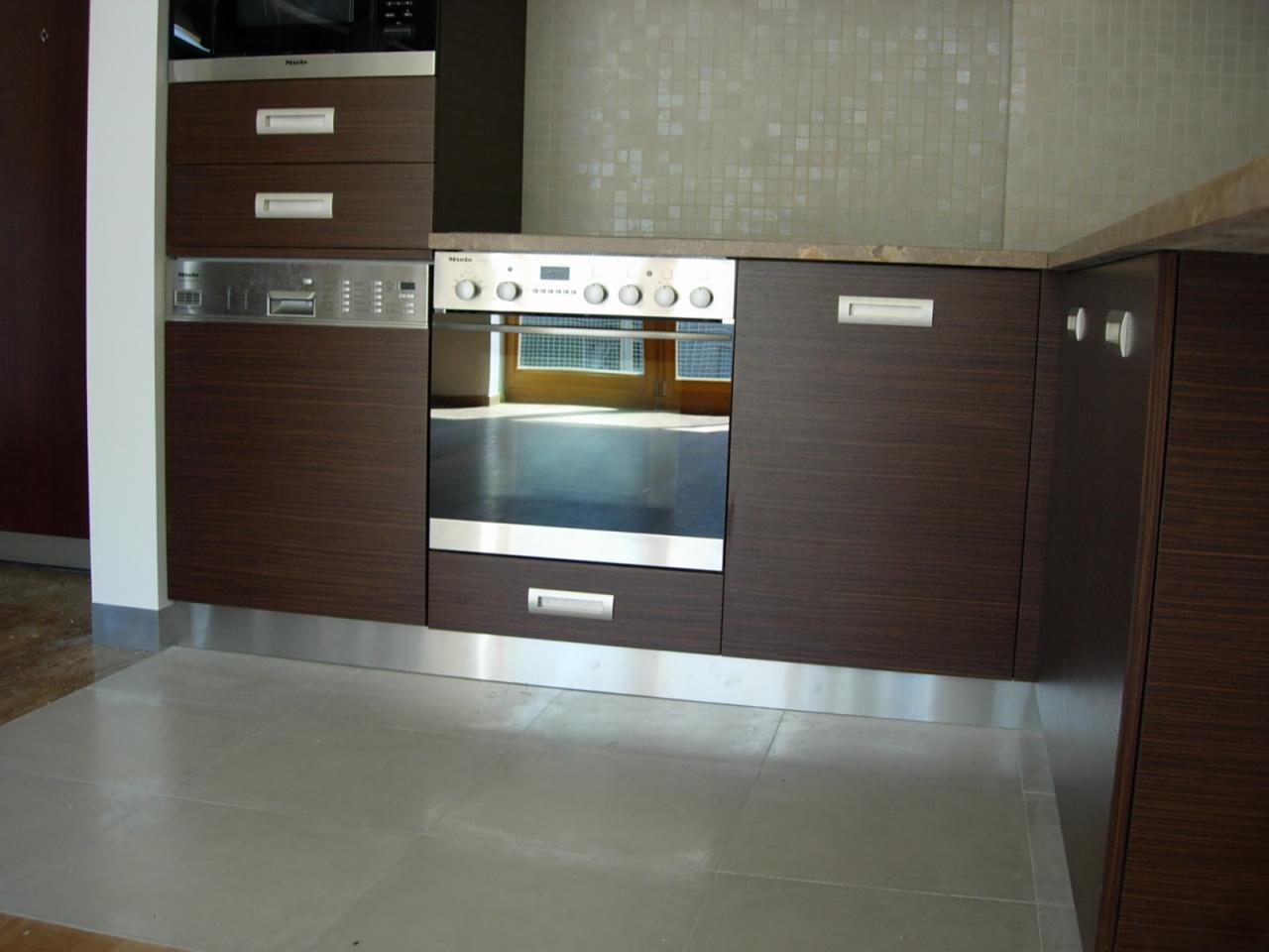 Nowoczesna zabudowa kuchenna z ciemnego drewna i stalowymi akcentami. Widoczny piekarnik oraz szuflady. Minimalistyczny design, jasne płytki na ścianie.