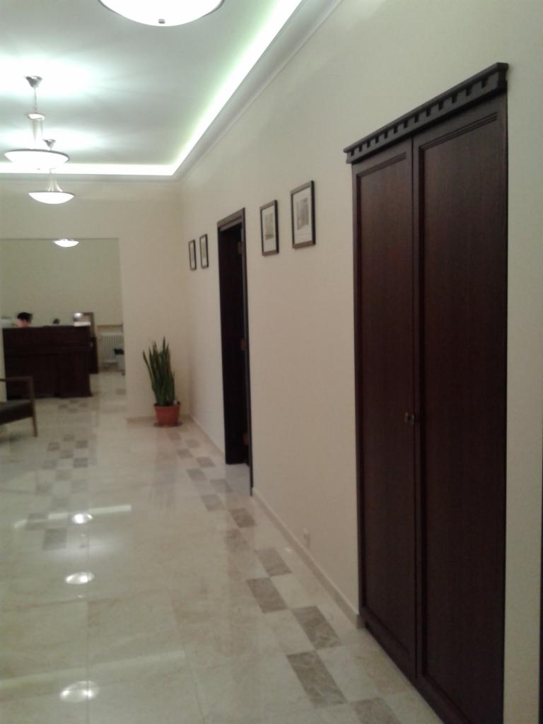 Elegancki korytarz z ciemnymi drzwiami i szafą wnękową. Podłoga z jasnych płytek w szachownicę. W tle recepcja i roślina doniczkowa. Oświetlenie sufitowe.