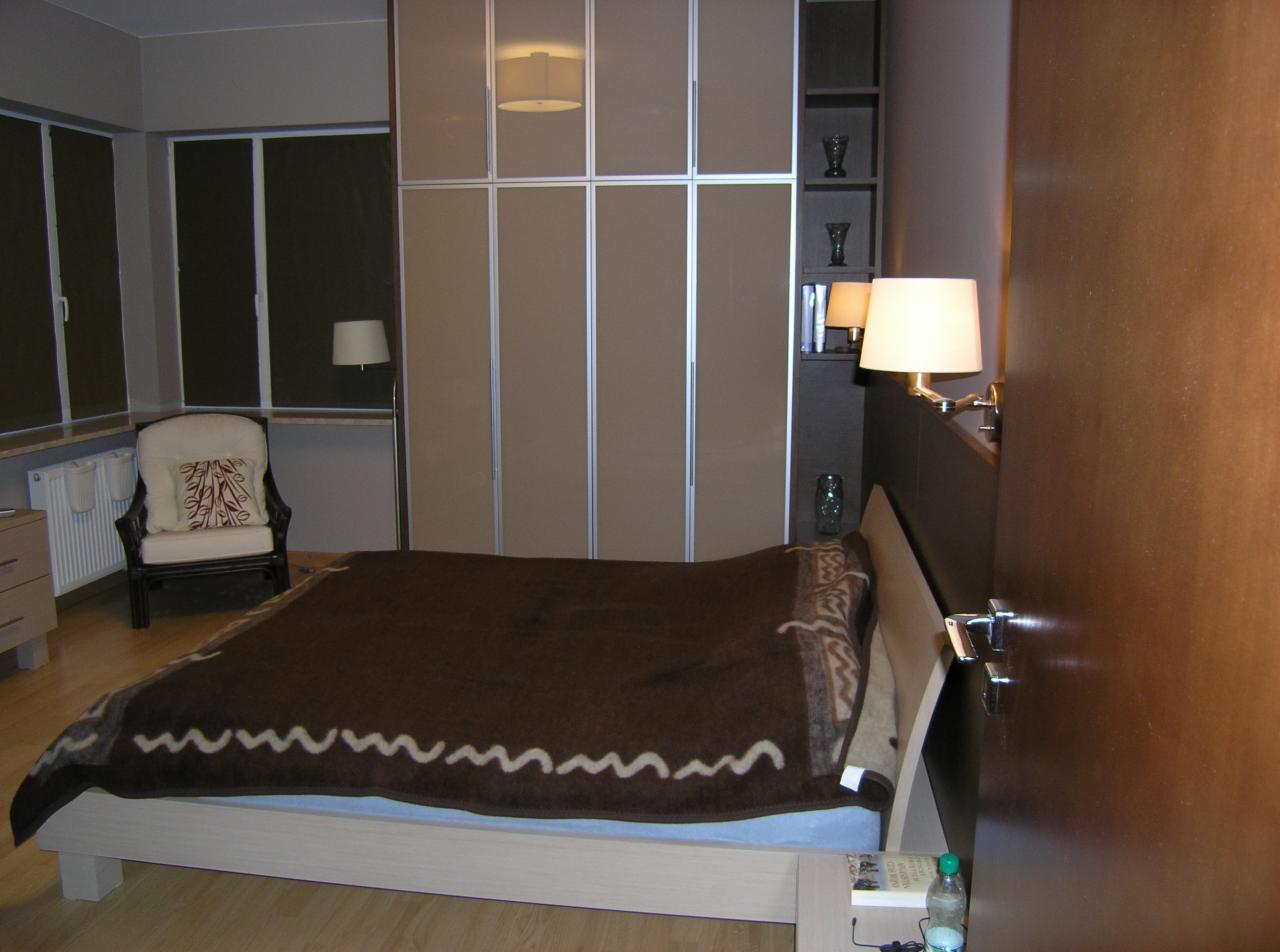 Sypialnia z dużą szafą wnękową z przesuwnymi drzwiami w kolorze beżowym, lampkami nocnymi i łóżkiem z brązową narzutą w pokoju z oknami i krzesłem.