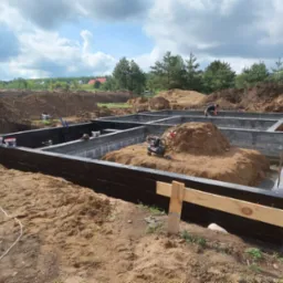 Wylane fundamenty pod budynek z izolacją przeciwwilgociową, widoczne szalunki, wykop, piasek, narzędzia i pracownik w tle.