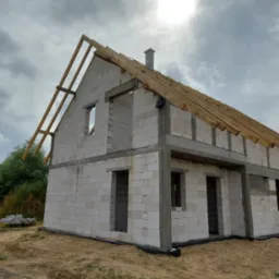 Surowy stan domu murowanego z białych bloczków, widoczna więźba dachowa w trakcie montażu, komin i niezabudowane okna na tle pochmurnego nieba.