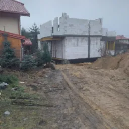 Wznoszenie ścian z białych bloczk&oacute;w na budowie domu jednorodzinnego, widoczne szalunki, teren wok&oacute;ł rozjeżdżony przez maszyny budowlane, pochmurne niebo.