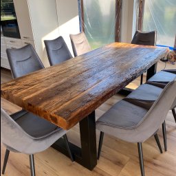 Jarosław Łyszcz - Stół z blatem z litego drewna o nieregularnych krawędziach, wsparty na czarnej, metalowej podstawie. Wokół stoją szare krzesła na cienkich, czarnych nogach.