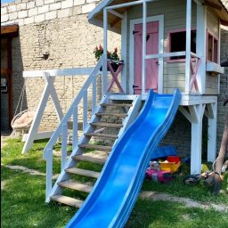 Jarosław Łyszcz - Drewniany domek dla dzieci z różowymi akcentami, schodami i zjeżdżalnią na trawniku, obok murowany budynek. Huśtawka w tle.