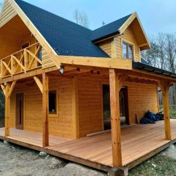 Jarosław Łyszcz - Drewniany dom z tarasem i balkonem, wykończony w naturalnym odcieniu drewna, z ciemnym dachem. Widok z zewnątrz, w tle drzewa.