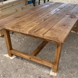 Jarosław Łyszcz - Rustykalny stół z drewna o ciepłym odcieniu, z widoczną strukturą słojów, ustawiony na zewnątrz. W tle stos desek, co sugeruje warsztat stolarski.