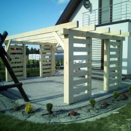 Jarosław Łyszcz - Surowa drewniana pergola w ogrodzie, widok z boku na konstrukcję z jasnego drewna, kostka brukowa, małe krzewy, huśtawka i dom w tle.