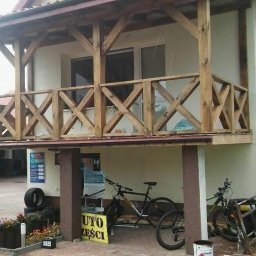 Jarosław Łyszcz - Drewniana konstrukcja balkonu z krzyżowymi balustradami, wsparta na kolumnach. W tle budynek z oknami i rynną. Widoczne rowery i drabina. Ujęcie z dołu, lekko z lewej strony.