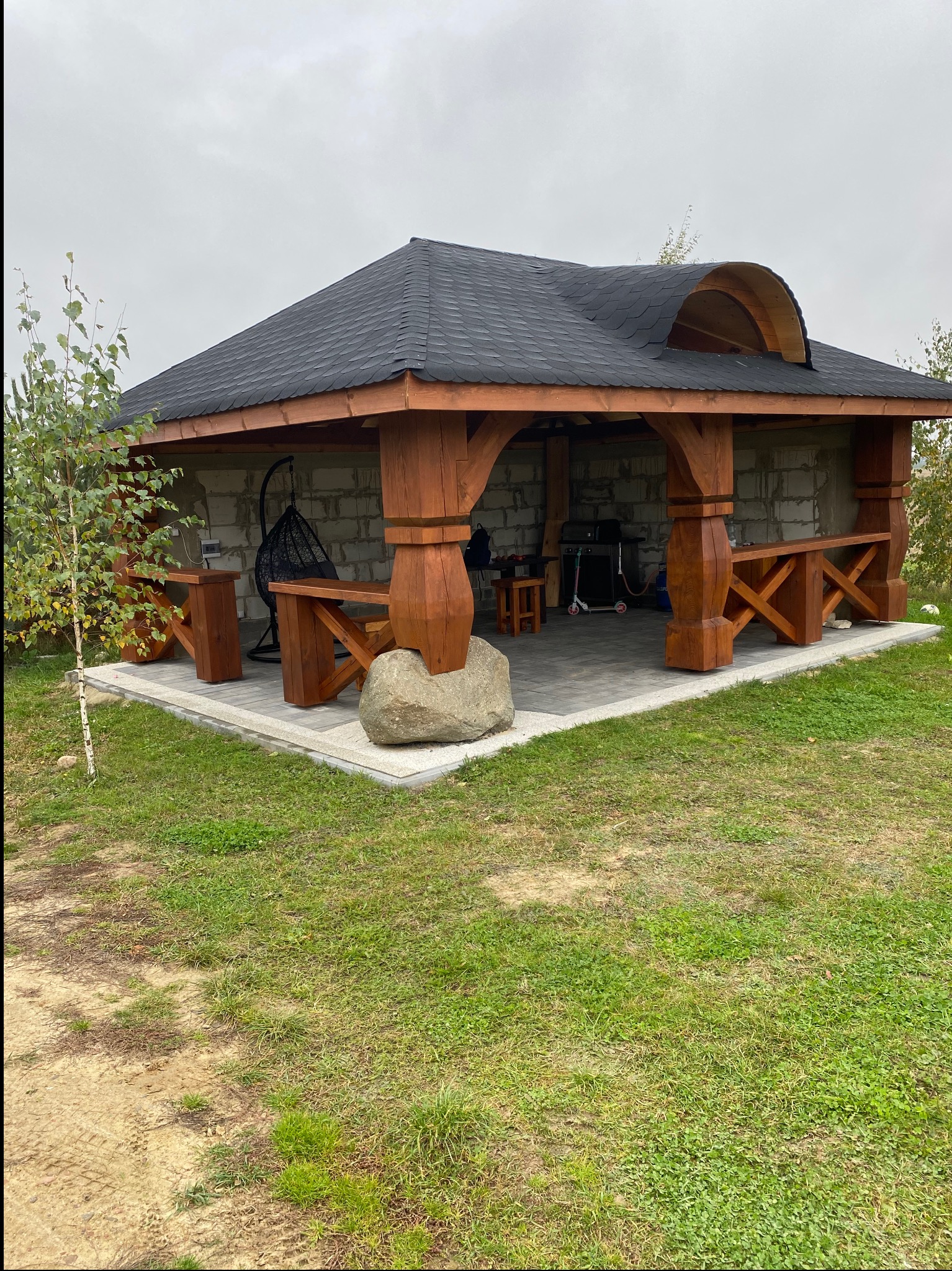 Zadaszona altana z ciemnym dachem i drewnianymi elementami konstrukcyjnymi. Wewnątrz grill i wiszące krzesło. Na zewnątrz trawnik i małe drzewko.