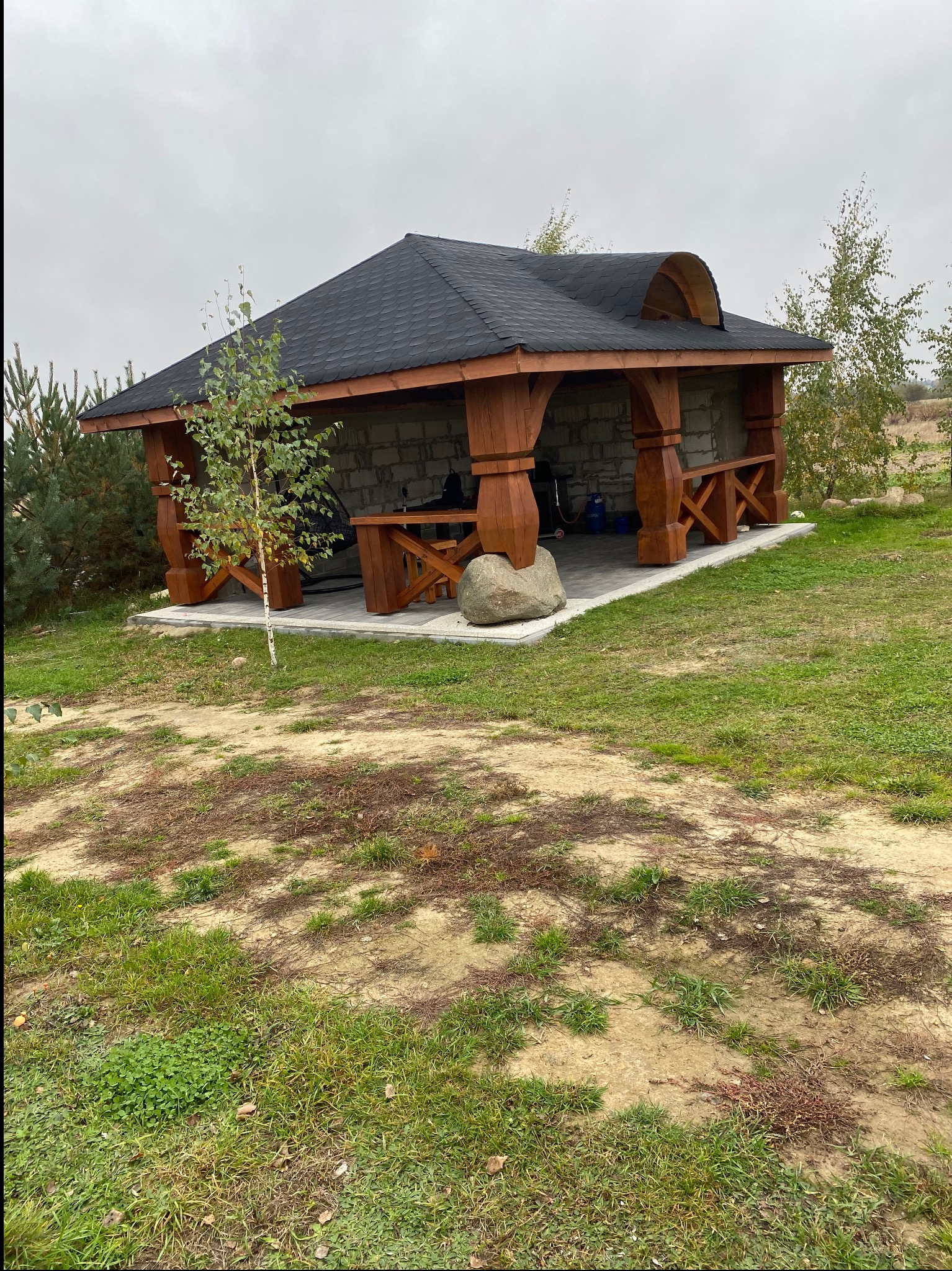 Drewniana altana z czarnym dachem, widoczna z zewnątrz, otoczona zielenią. Elementy konstrukcyjne z brązowego drewna, ściany z szarego bloczka. Na pierwszym planie mała brzoza.