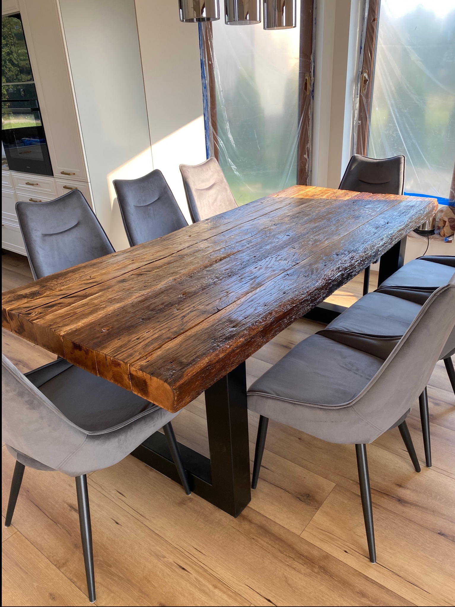 Stół z blatem z litego drewna o nieregularnych krawędziach, wsparty na czarnej, metalowej podstawie. Wokół stoją szare krzesła na cienkich, czarnych nogach.