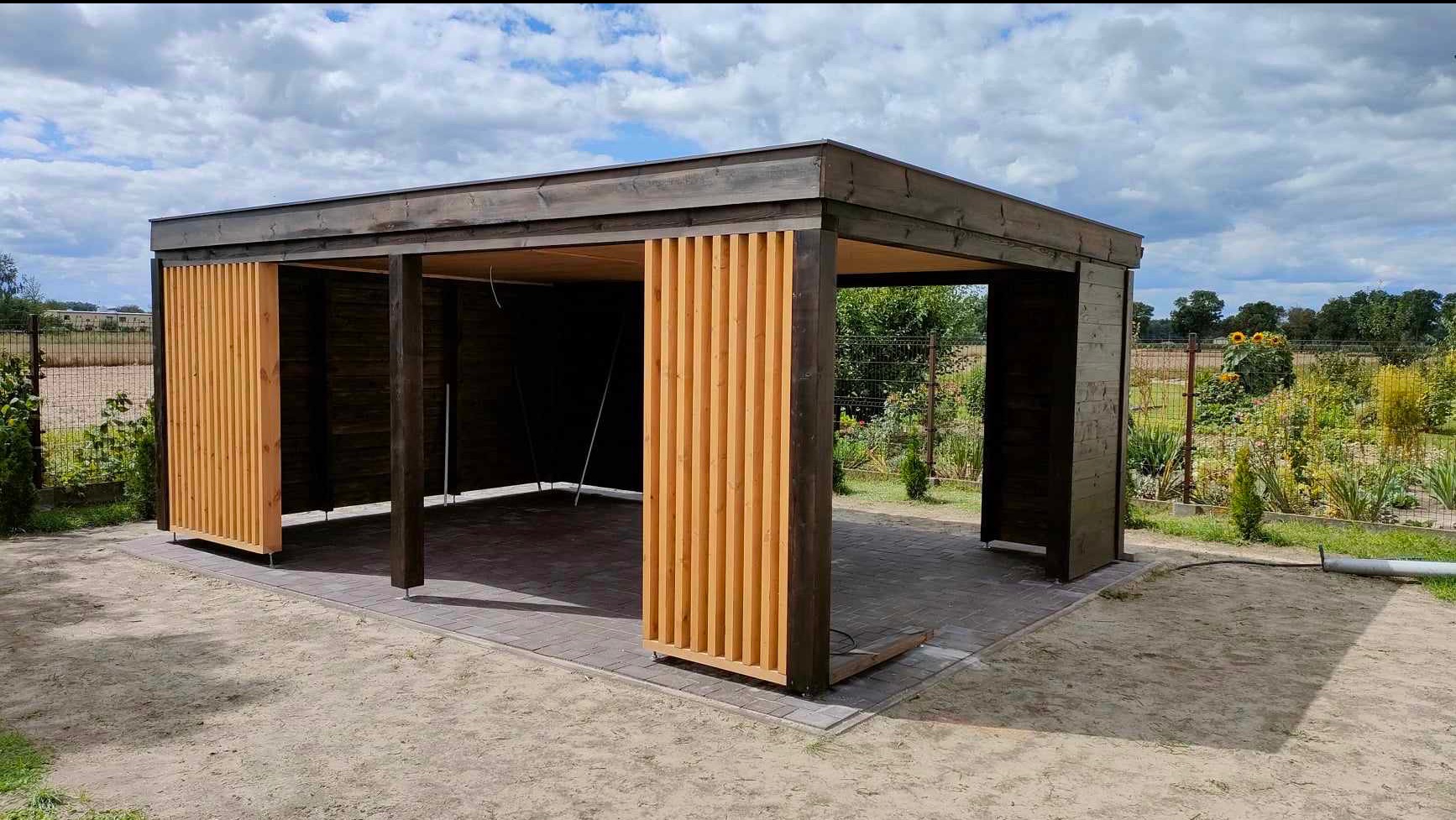 Nowoczesna altana z ciemnego drewna z jasnymi, pionowymi panelami, wykończona kostką brukową. Widok na ogród z zielenią i słonecznikami w tle.