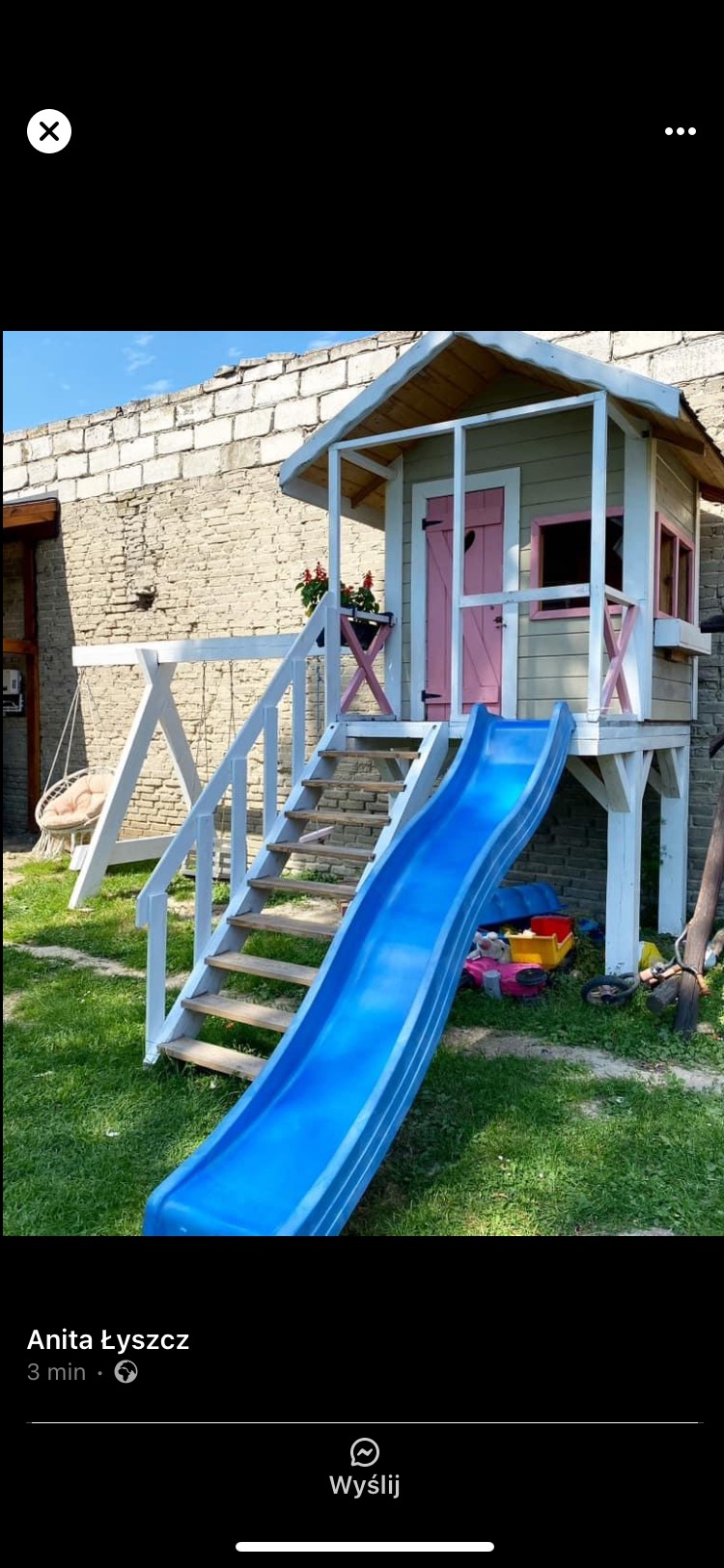 Drewniany domek dla dzieci z różowymi akcentami, schodami i zjeżdżalnią na trawniku, obok murowany budynek. Huśtawka w tle.