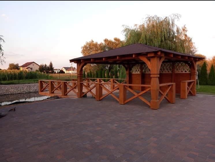 Drewniana altana z brązowym dachem, połączona z tarasem i małym mostkiem nad wodą. Całość w otoczeniu zieleni i domów.