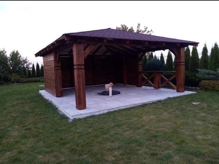 Drewniana altana z ciemnym dachem na jasnej, kamiennej podłodze. Widoczne zdobienia na słupach i krzyżowe elementy balustrady. Tło stanowi zielony trawnik i wysokie tuje.