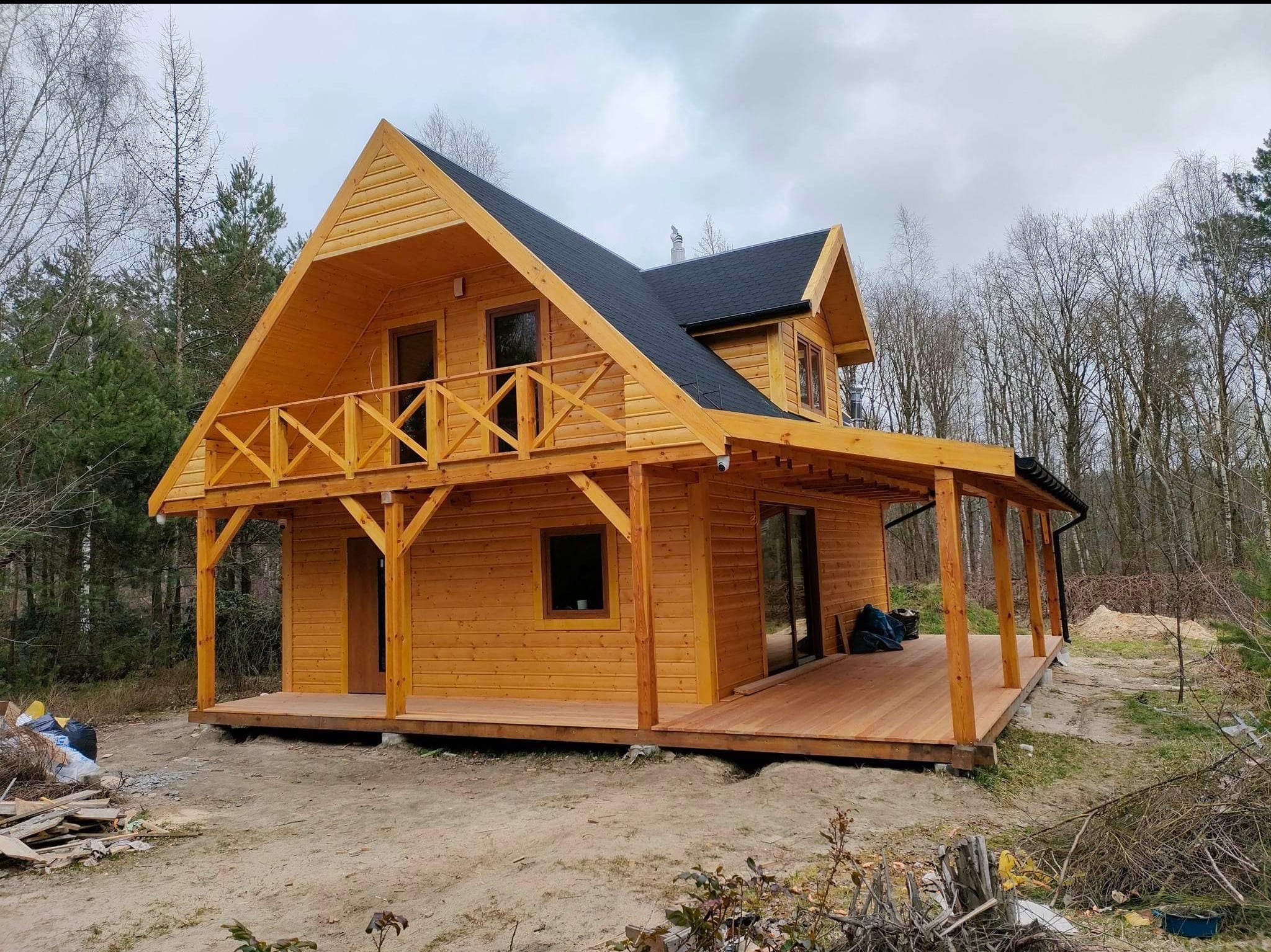 Drewniany dom z balkonem i tarasem, wykończony w naturalnym kolorze drewna, na tle leśnym. Widoczne detale konstrukcyjne i czarne pokrycie dachu.