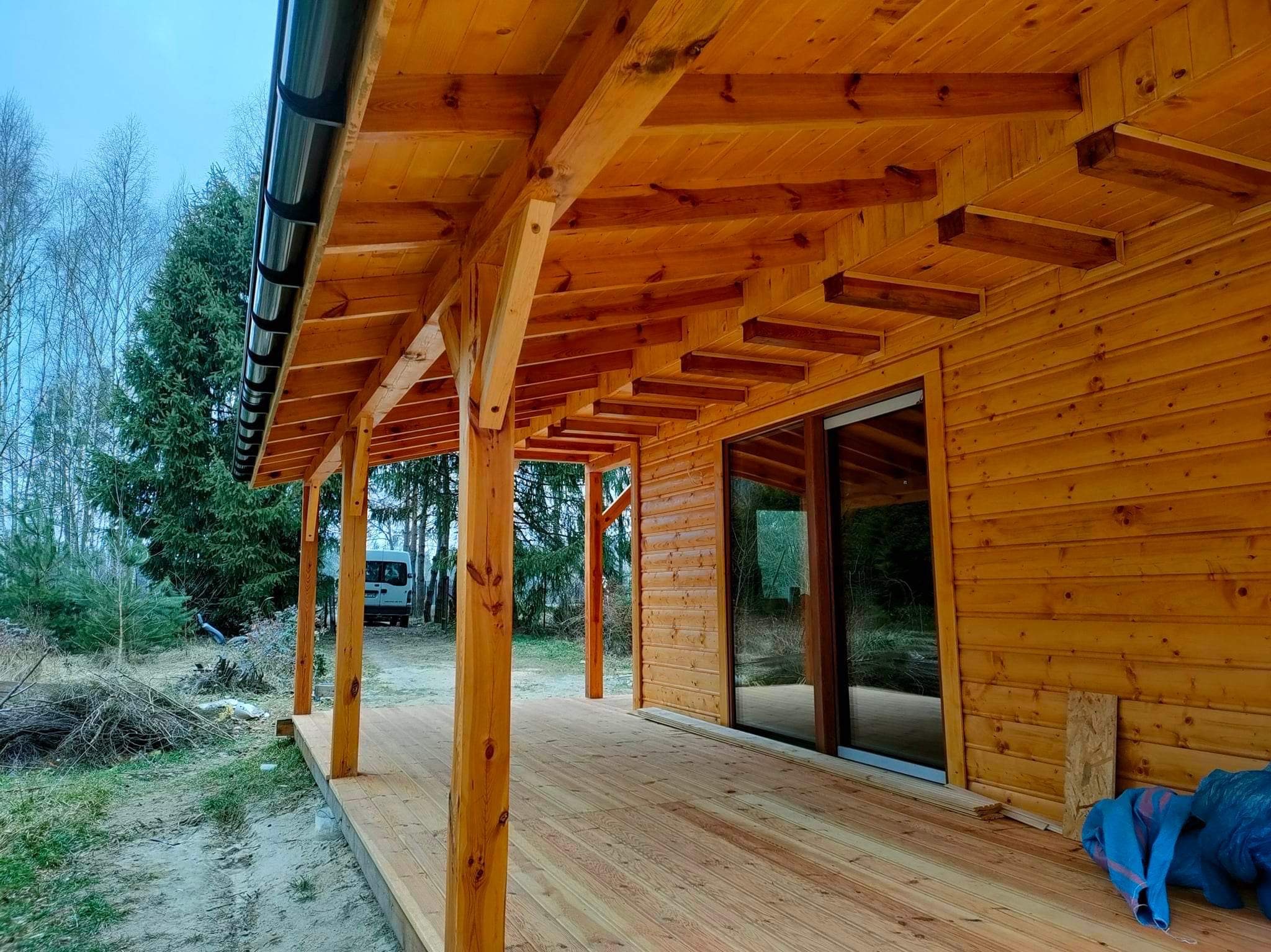 Drewniana altana z zadaszeniem i tarasem, widok z zewnątrz. Konstrukcja z belek, widoczne łączenia. W tle drzewa i częściowo widoczny samochód dostawczy.