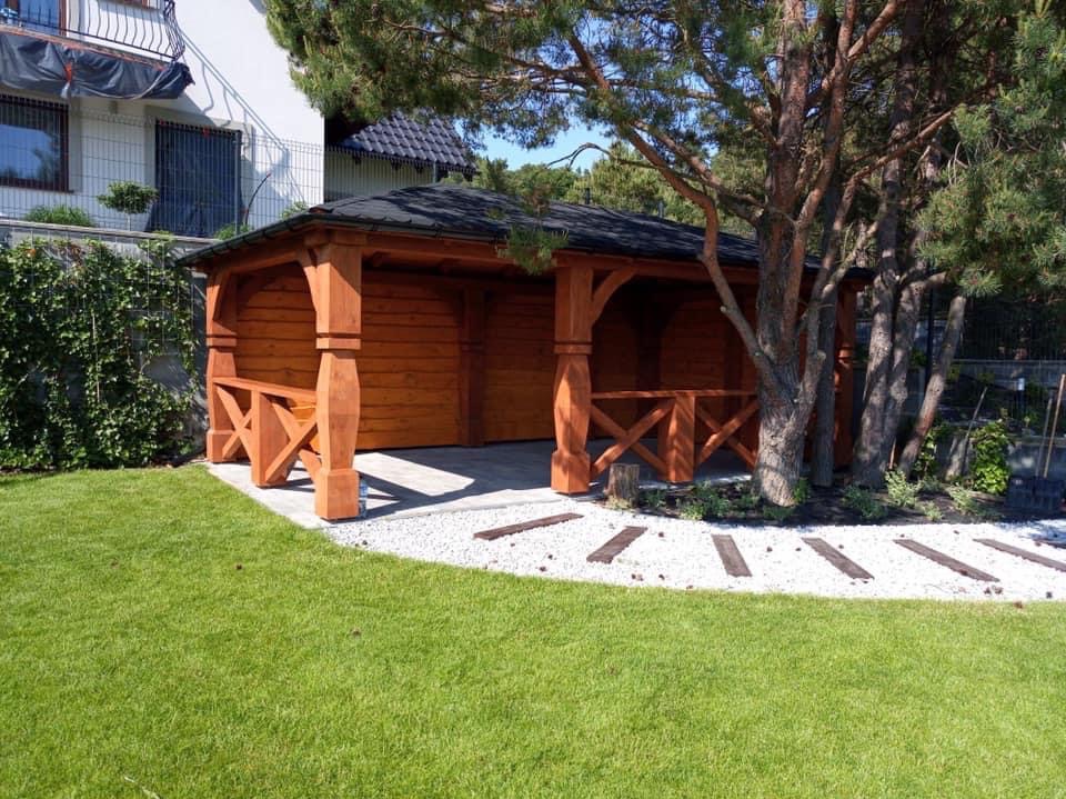 Drewniana altana z czarnym dachem, otoczona zielonym trawnikiem i białym żwirem z drewnianymi elementami. Widoczne drzewa i budynek w tle.
