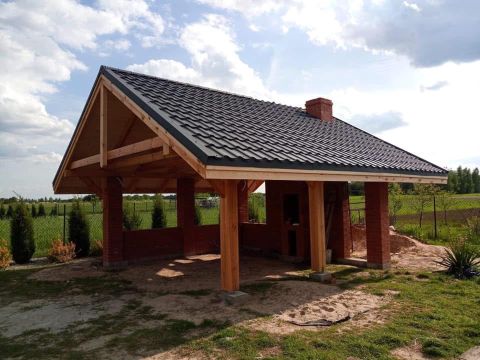 Murowana altana z drewnianą konstrukcją dachu krytego ciemną dachówką, z kominem, na tle zieleni i błękitnego nieba z chmurami.