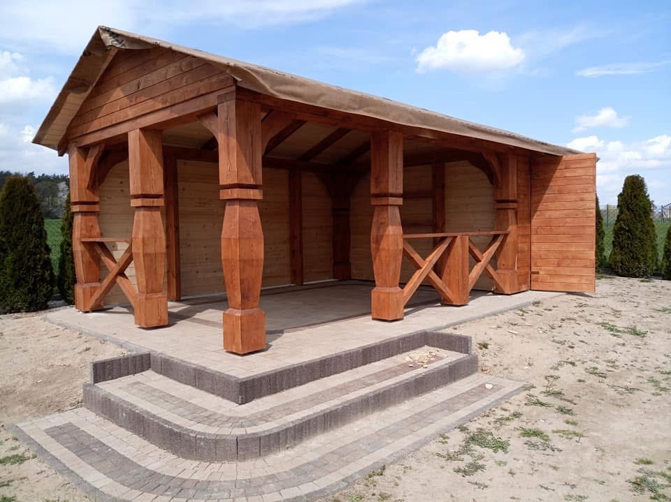 Drewniana altana z beżowym zadaszeniem, widoczne zdobione słupy i schody z kostki brukowej. Całość w otoczeniu zieleni i błękitnego nieba.