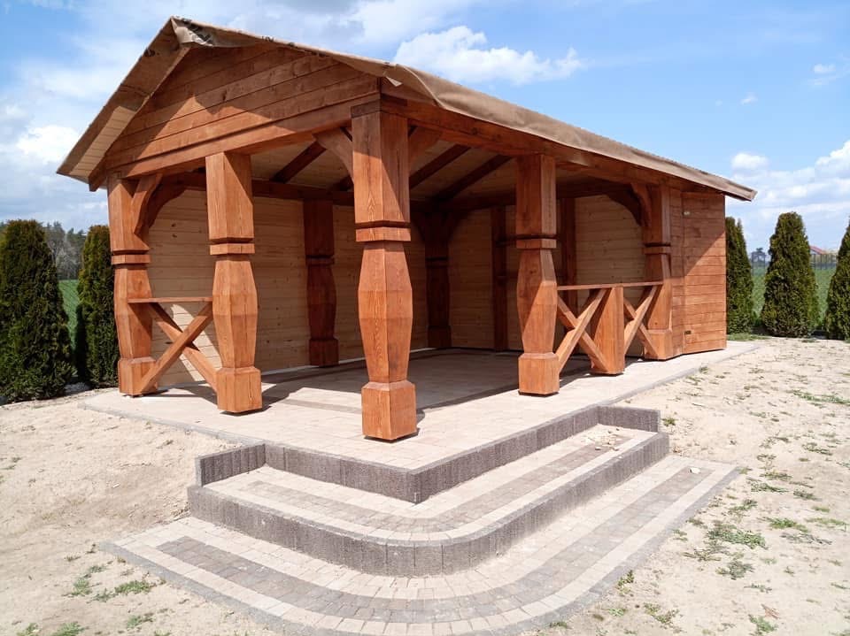 Drewniana altana z czterema schodkami wejściowymi wyłożonymi kostką brukową, otoczona tujami i piaskiem, pod błękitnym niebem.