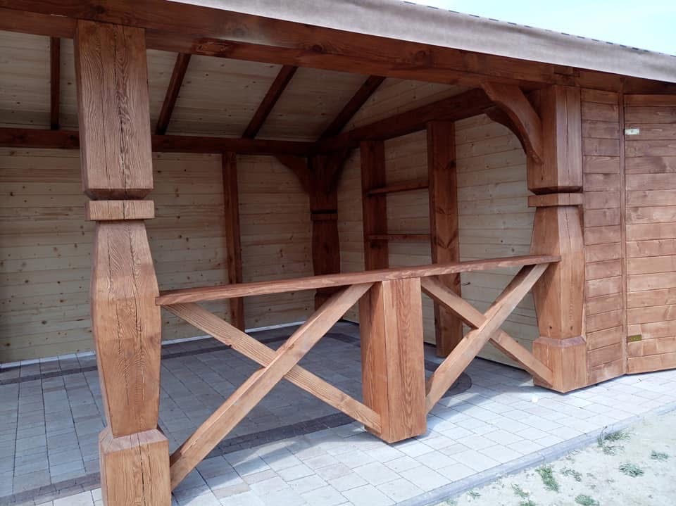 Drewniana altana z balustradą i widoczną konstrukcją dachu, wykończona w ciepłym odcieniu brązu, na tle jasnej, drewnianej ściany i kostki brukowej.