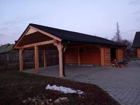 Drewniana konstrukcja z zadaszeniem i budynkiem z bali w tle, widok z zewnątrz. Elementy drewniane w naturalnym kolorze, czarny dach. Ułożona kostka brukowa.