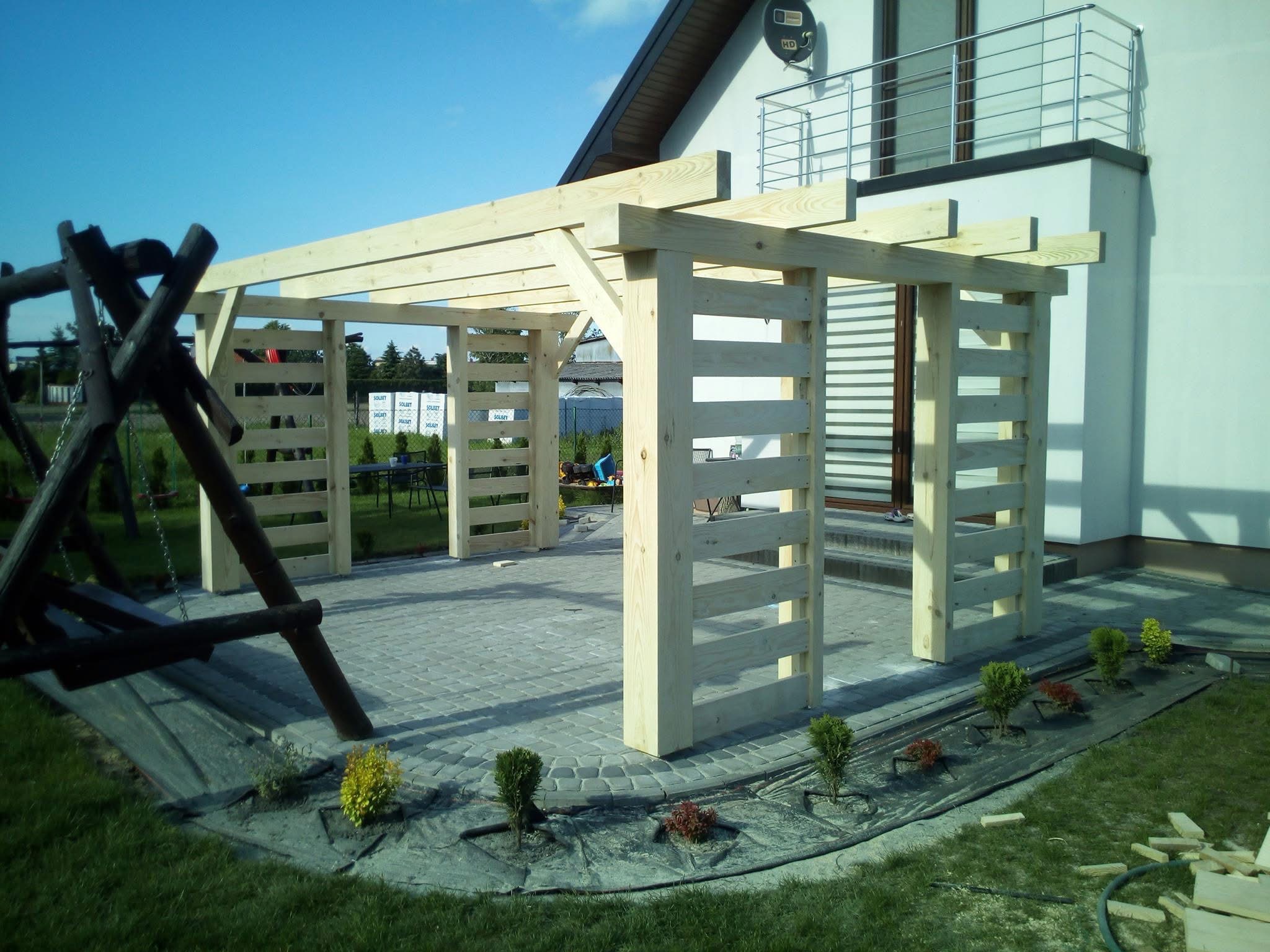 Surowa drewniana pergola w ogrodzie, widok z boku na konstrukcję z jasnego drewna, kostka brukowa, małe krzewy, huśtawka i dom w tle.