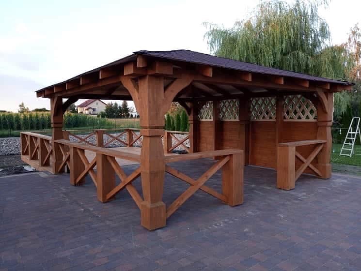 Drewniana altana z brązowym dachem na kostce brukowej. Widoczne ozdobne kraty i barierki. W tle zieleń i dom. Drabina oparta o ścianę altany.