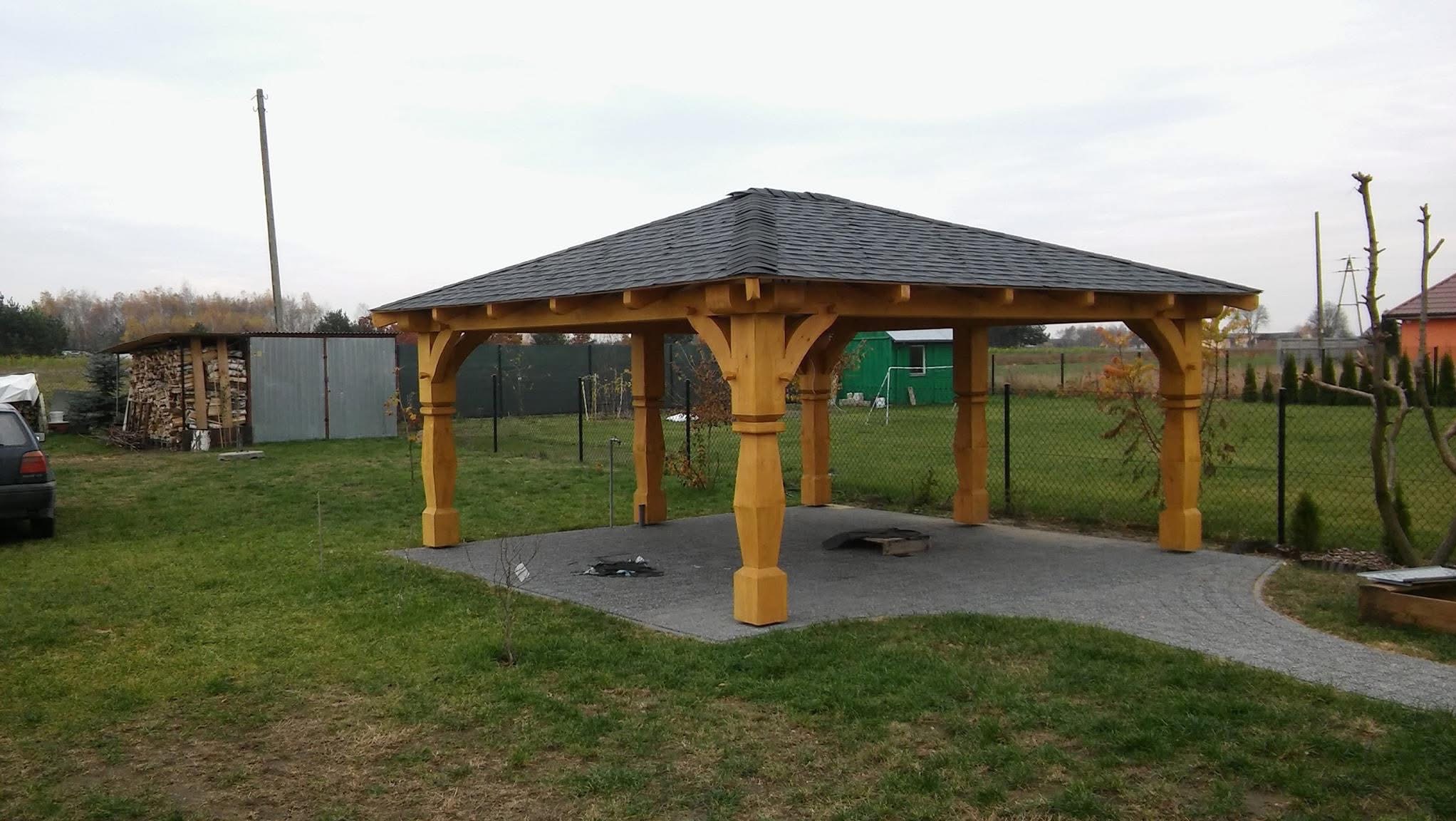 Drewniana altana z ciemnym dachem na betonowej podstawie, otoczona zielonym trawnikiem i ogrodzeniem z siatki. W tle widoczne drzewa i budynki.