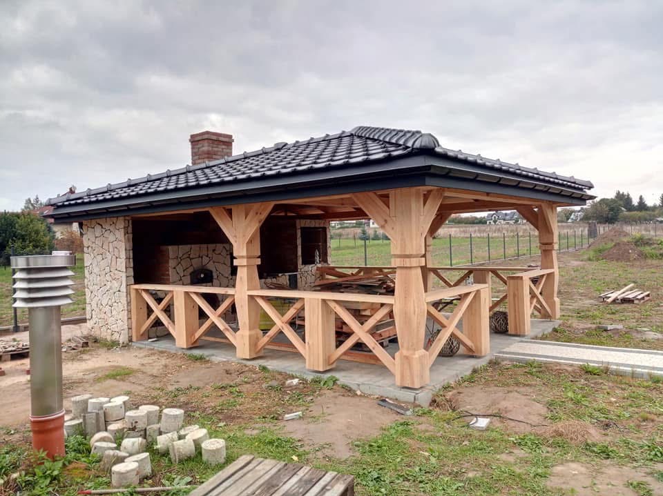 Drewniana altana z kamiennym grillem i czarnym dachem z dachówki, otoczona zielenią. Widoczne elementy konstrukcyjne i wykończeniowe. Częściowo ukończona budowa.