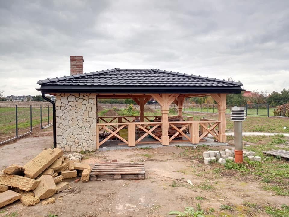 Kamienna altana z drewnianą konstrukcją i ciemnym dachem, widoczna z zewnątrz, z materiałami budowlanymi obok. Ujęcie w pochmurny dzień.