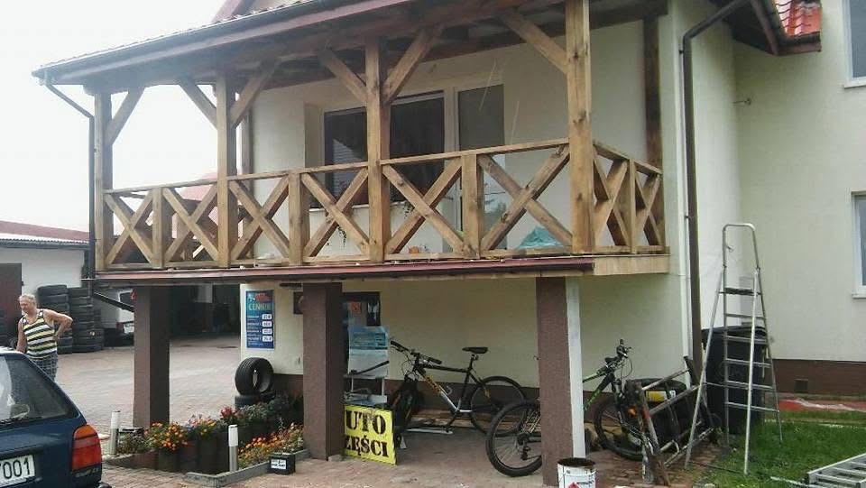 Drewniana konstrukcja balkonu z krzyżowymi balustradami, wsparta na kolumnach. W tle budynek z oknami i rynną. Widoczne rowery i drabina. Ujęcie z dołu, lekko z lewej strony.