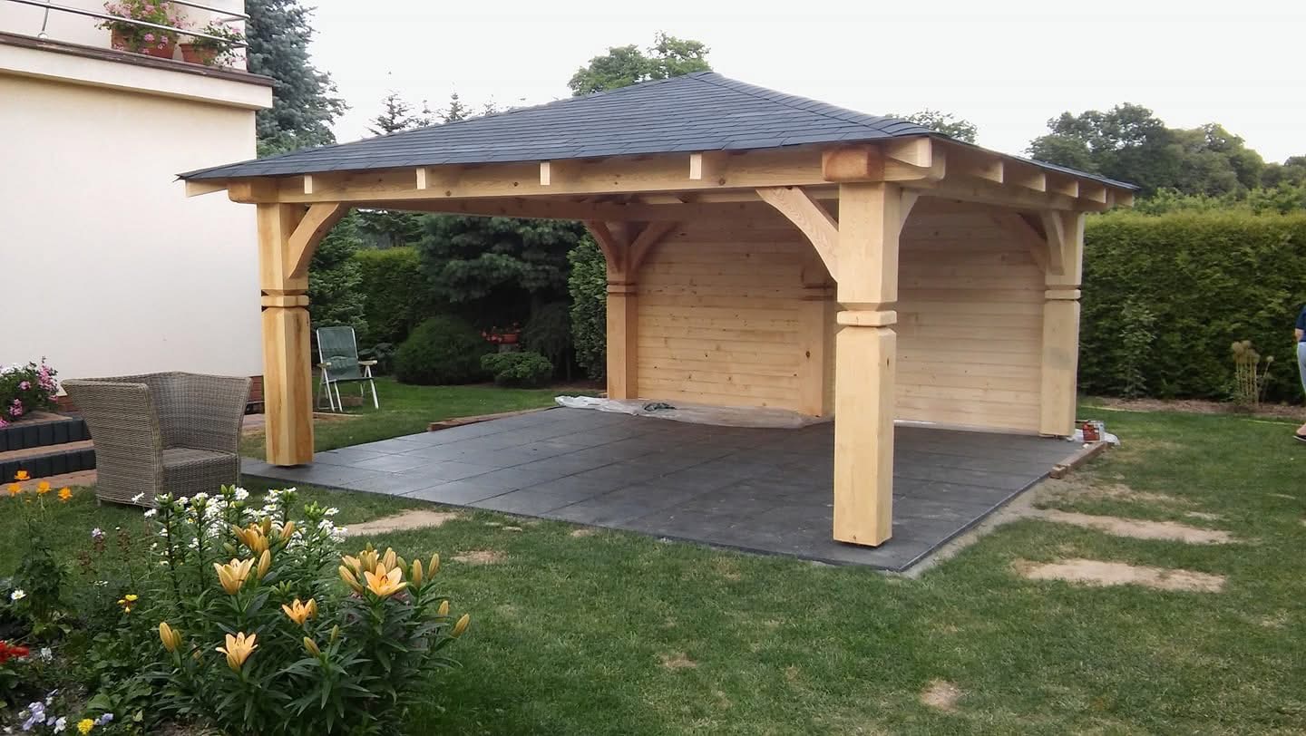 Drewniana altana z ciemnym dachem na betonowej podstawie, otoczona zielenią. W tle budynek i wysokie drzewa. W pobliżu kwiaty i wiklinowy fotel.