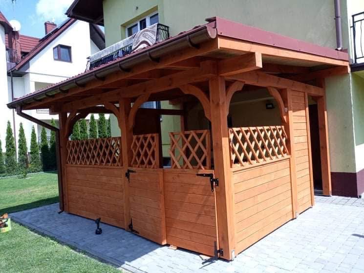 Drewniana altana z kratownicą i zadaszeniem w kolorze ceglastym, umieszczona na kostce brukowej, z widocznym fragmentem budynku w tle.