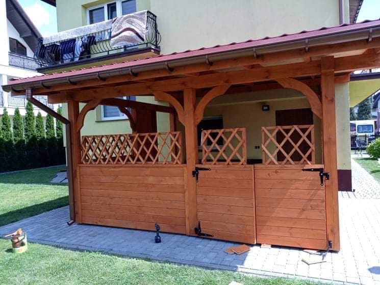 Drewniana altana z kratką i zamykanymi drzwiami, pomalowana na brązowo, z widocznym wiaderkiem farby i pędzlem, na tle domu z balkonem i zielonego trawnika.