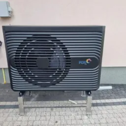 Czarna jednostka zewnętrzna z poziomymi paskami i logo FOX Air na metalowej podstawie, zamontowana przy jasnej ścianie budynku z widoczną rurą spustową i skrzynką elektryczną.