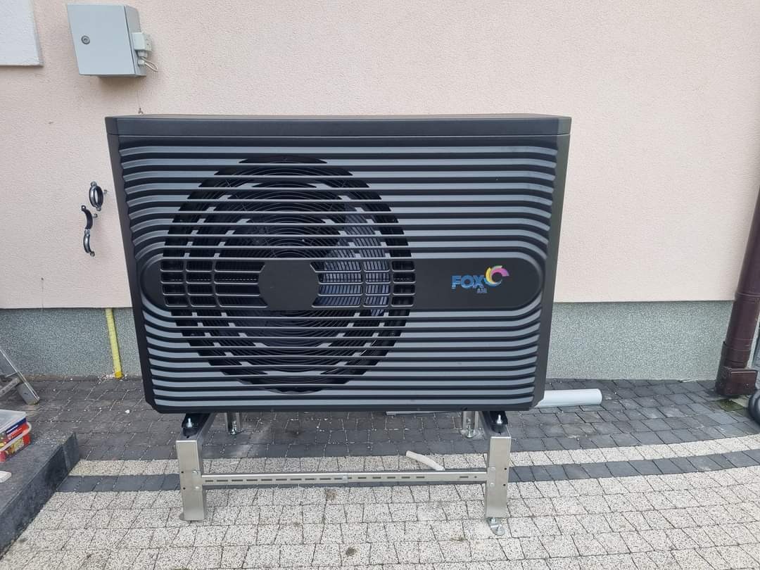 Czarna jednostka zewnętrzna z poziomymi paskami i logo FOX Air na metalowej podstawie, zamontowana przy jasnej ścianie budynku z widoczną rurą spustową i skrzynką elektryczną.