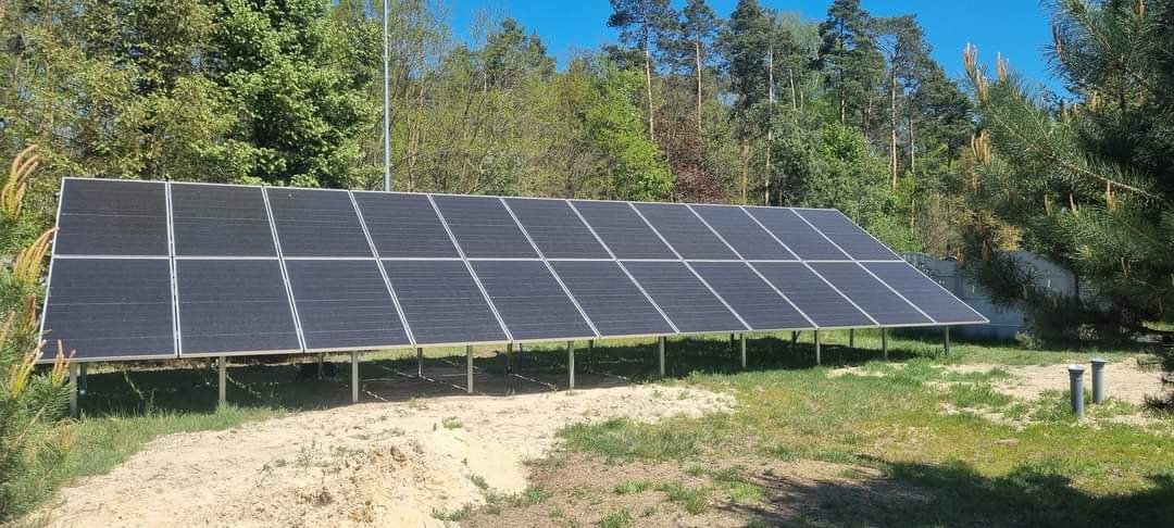 Rozległa instalacja paneli słonecznych na metalowej konstrukcji, umieszczona na trawiastym terenie z piaszczystym podłożem, otoczona drzewami w słoneczny dzień.