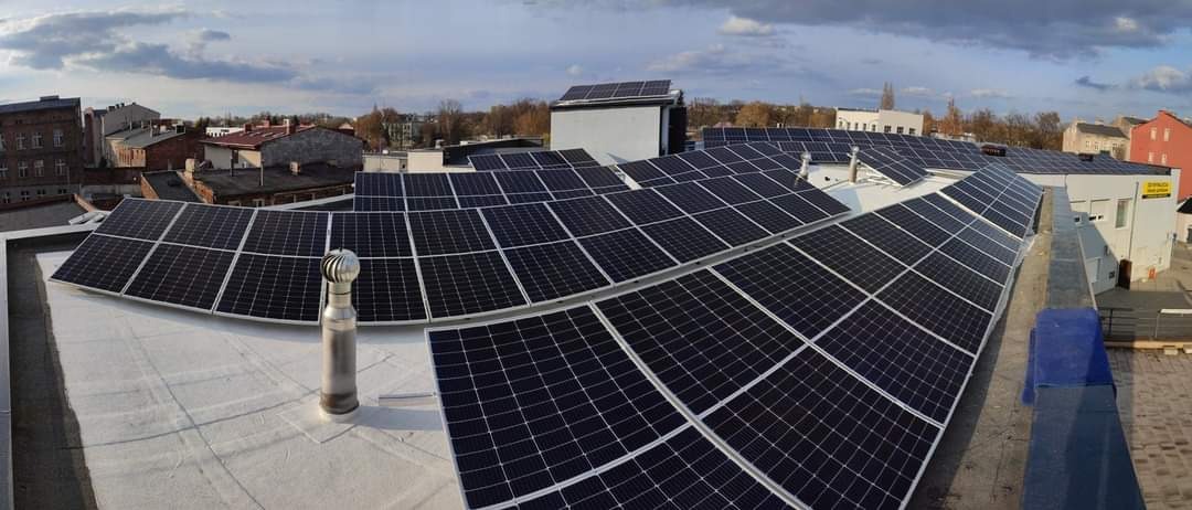 Rozległa instalacja paneli słonecznych na płaskim dachu budynku w miejskiej scenerii, widoczny komin wentylacyjny i budynki w tle pod pochmurnym niebem.